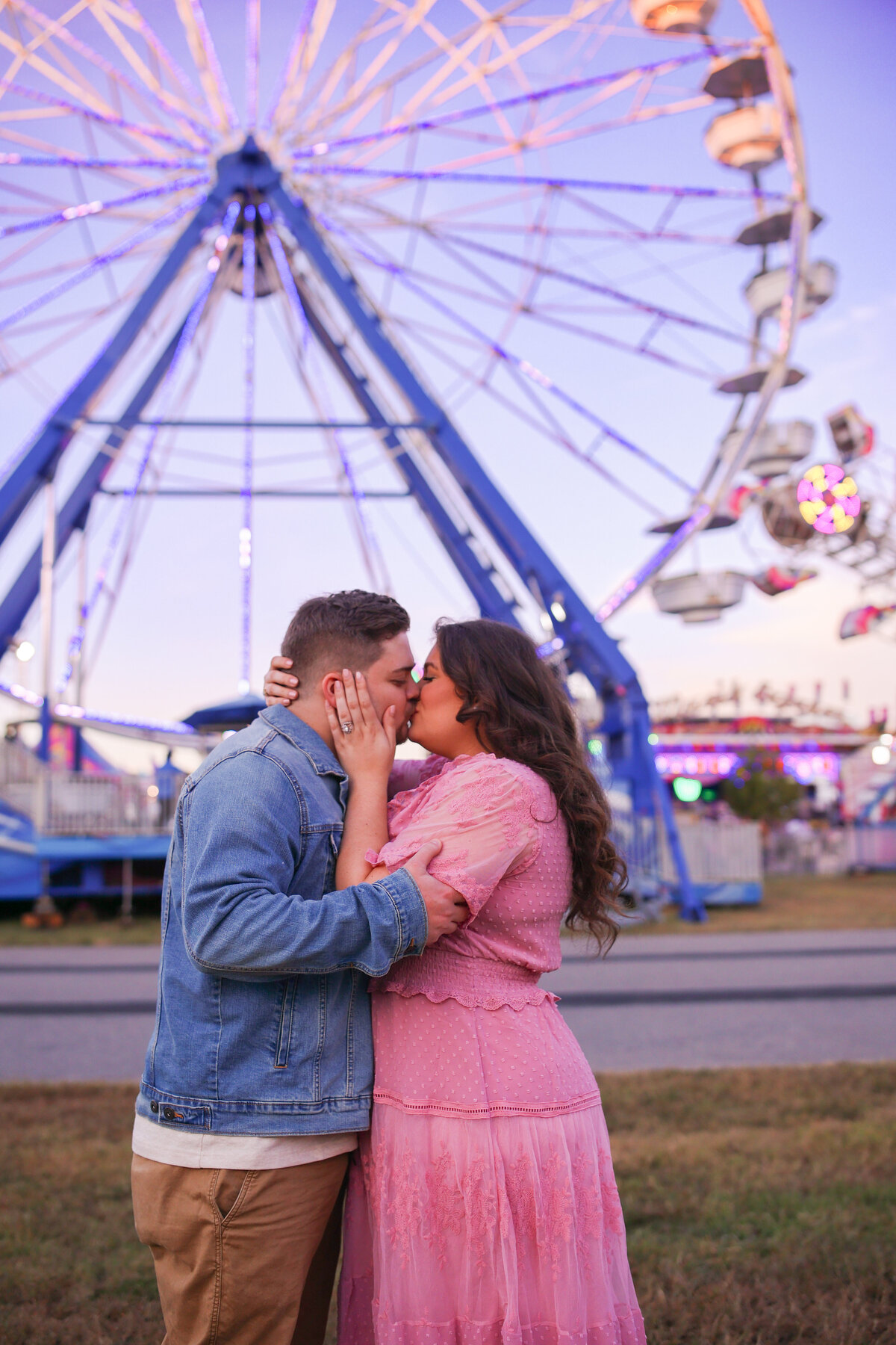 man and woman kissing with a ferris wheel in the background at sunset
