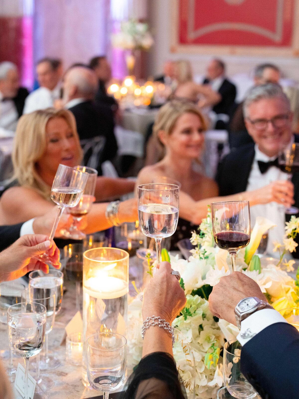 A group of elegantly dressed people raise their glasses in a toast around a table adorned with candles and floral arrangements at a formal event. The atmosphere is lively and celebratory.