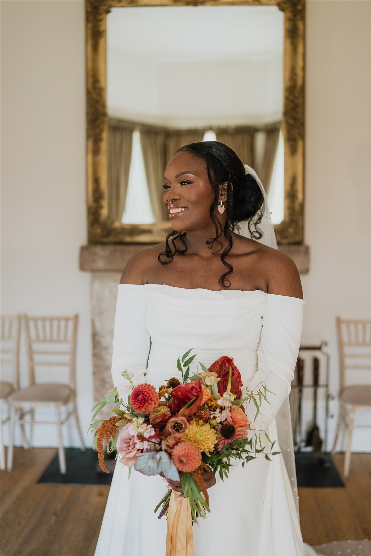 A bride wears a long white wedding dress and is photographed holding her bouquet in the empty dining room at Logie Country House. A large mirror hangs behind her and she smiles candidly.