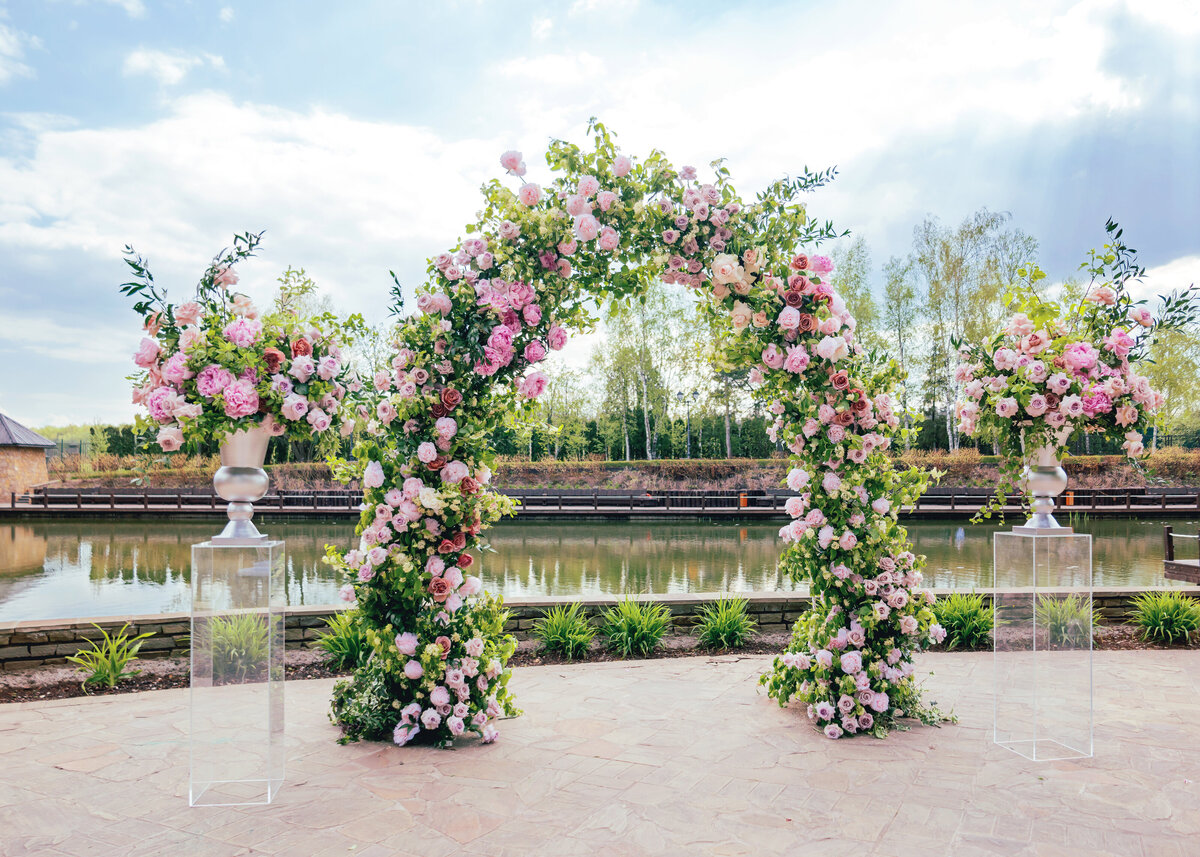 Beautiful floral arch for wedding ceremony. Vases with pink roses and peonies. Wedding set up outdoors in park near lake