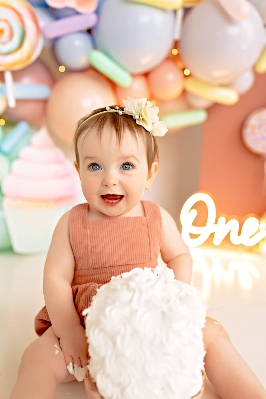 A smiling baby wearing a peach-colored outfit and a floral headband sits in front of colorful balloons and a light-up sign reading "One." The baby is holding a cake with white frosting, signifying a first birthday celebration.