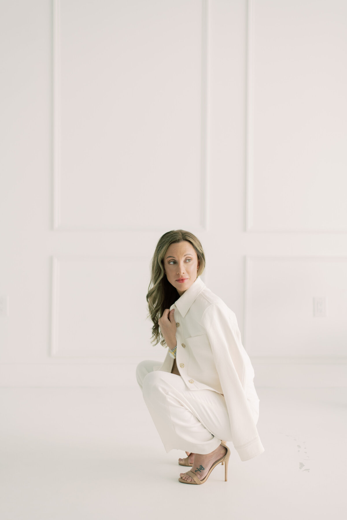 Empowered studio portrait of woman crouched in all-white suit and heels, direct gaze
