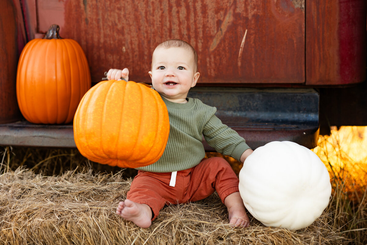 Baby boy lifts fake pumpkin in the air and smiles.