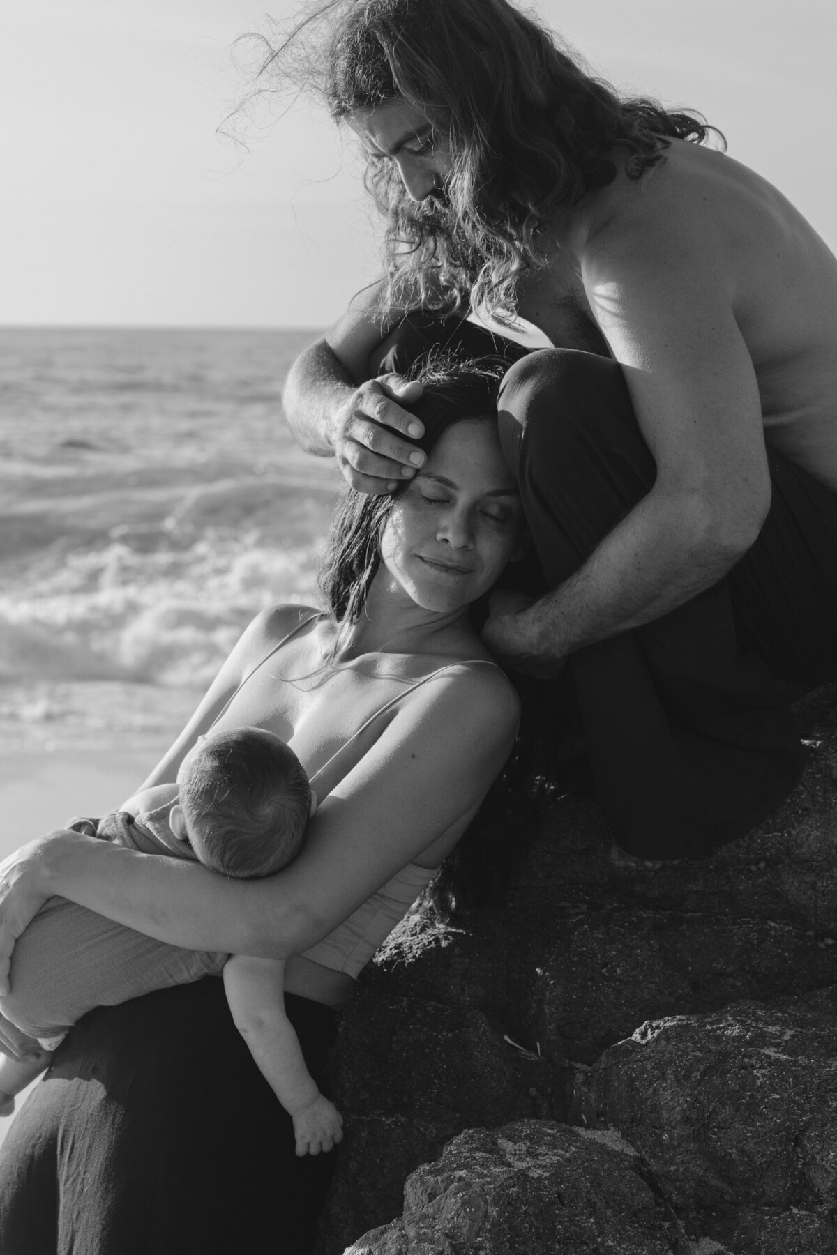 Black and white beach scene featuring a mother breastfeeding her newborn while the father tenderly supports her, captured by Karen Pride