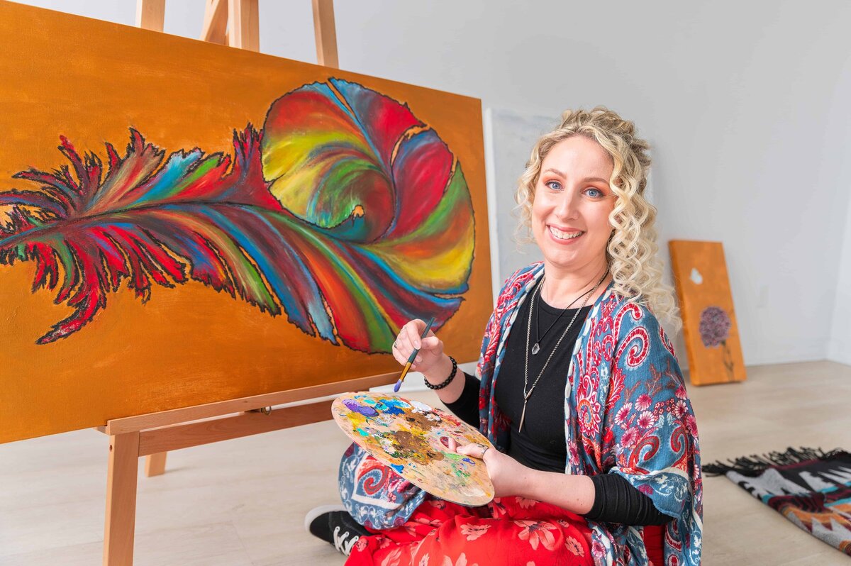 Client smiling for camera while seated next to their canvas with palette in hand