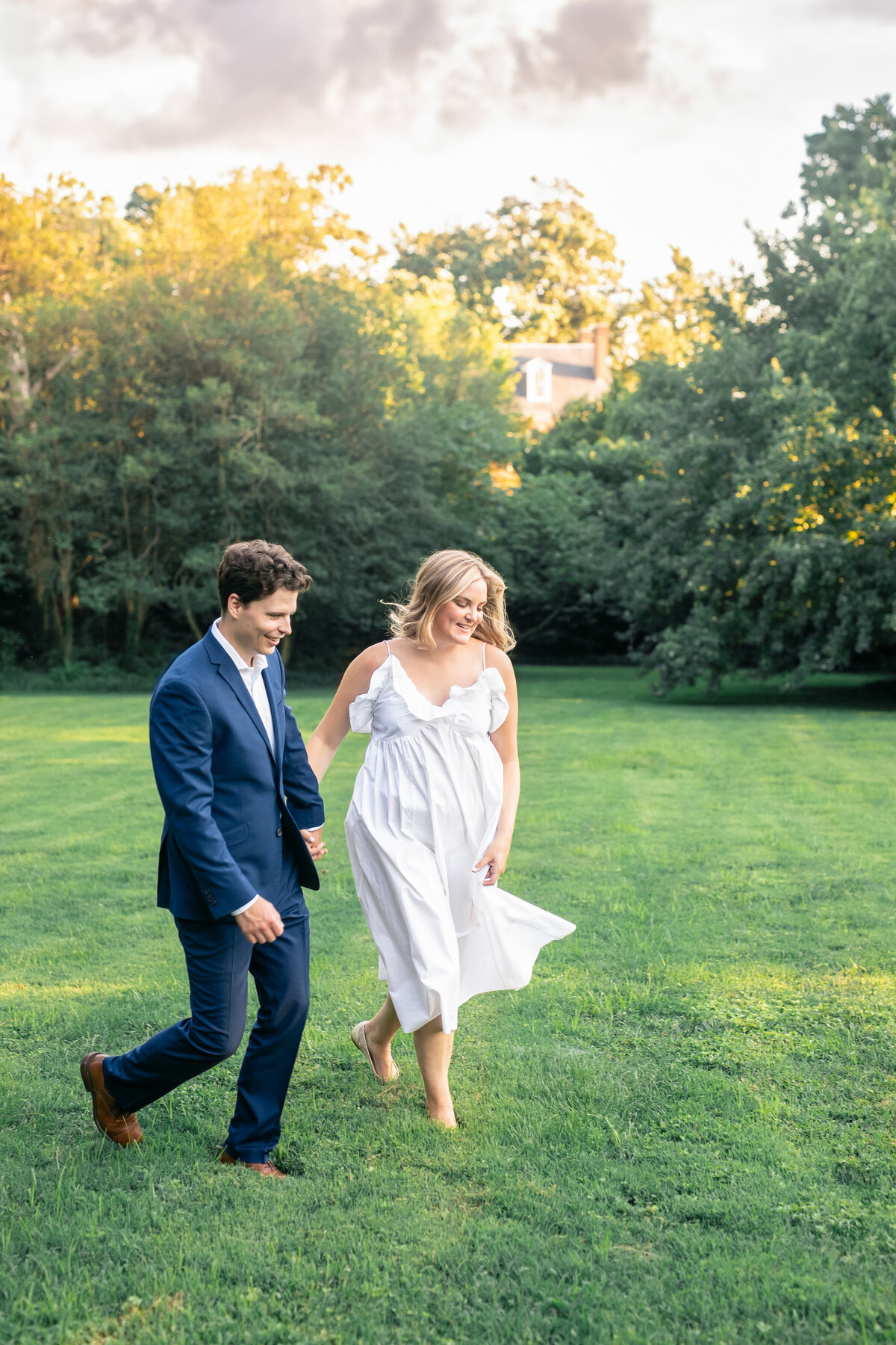 A couple walks hand in hand on a grassy field. The man is wearing a blue suit, and the woman is in a flowing white dress. The sun sets behind trees in the background, creating a warm and romantic atmosphere.
