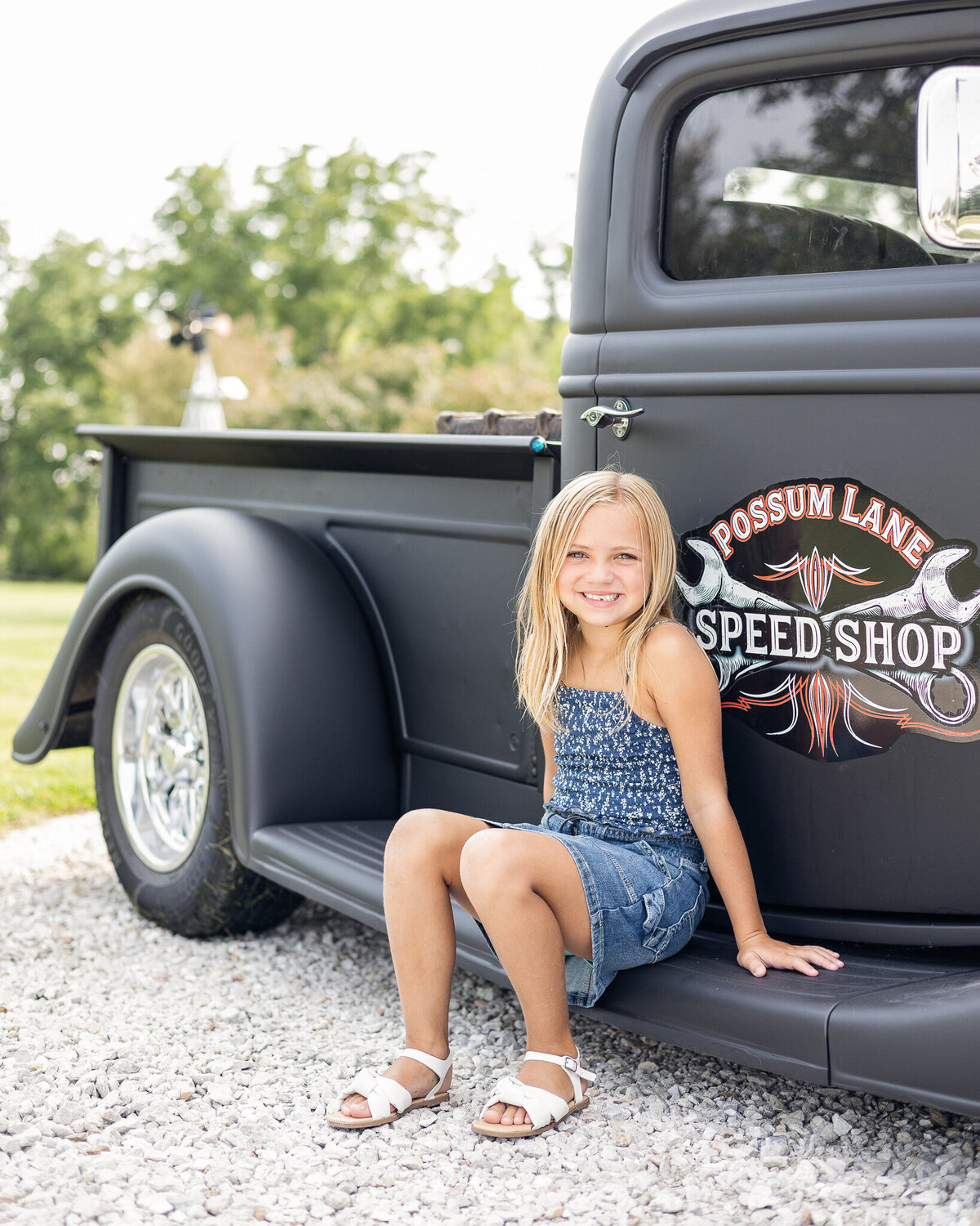Youngest daughter with medium length blonde hair, wearing a blue jean skirt, dark white and blue tank style shirt, white shoes, and smiling at the camera.  She's sitting on the running board of the black classic car in the rocked drive.