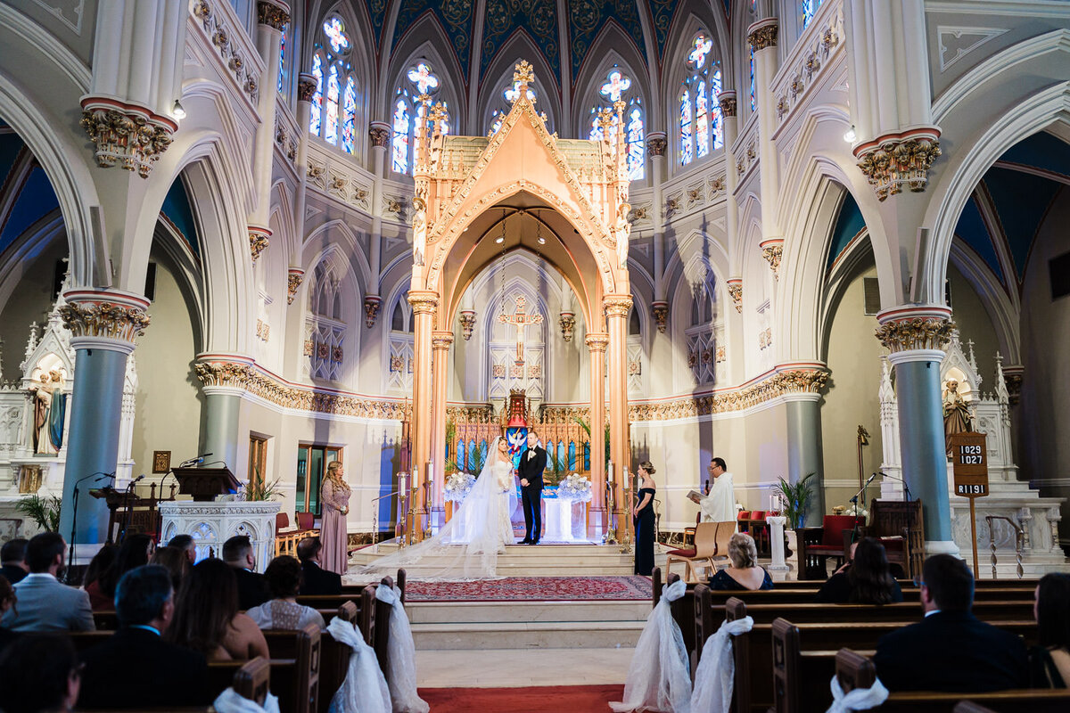 Bride and groom standing at the altar of a grand cathedral during their wedding ceremony, with ornate arches, stained-glass windows, and guests seated in the pews.