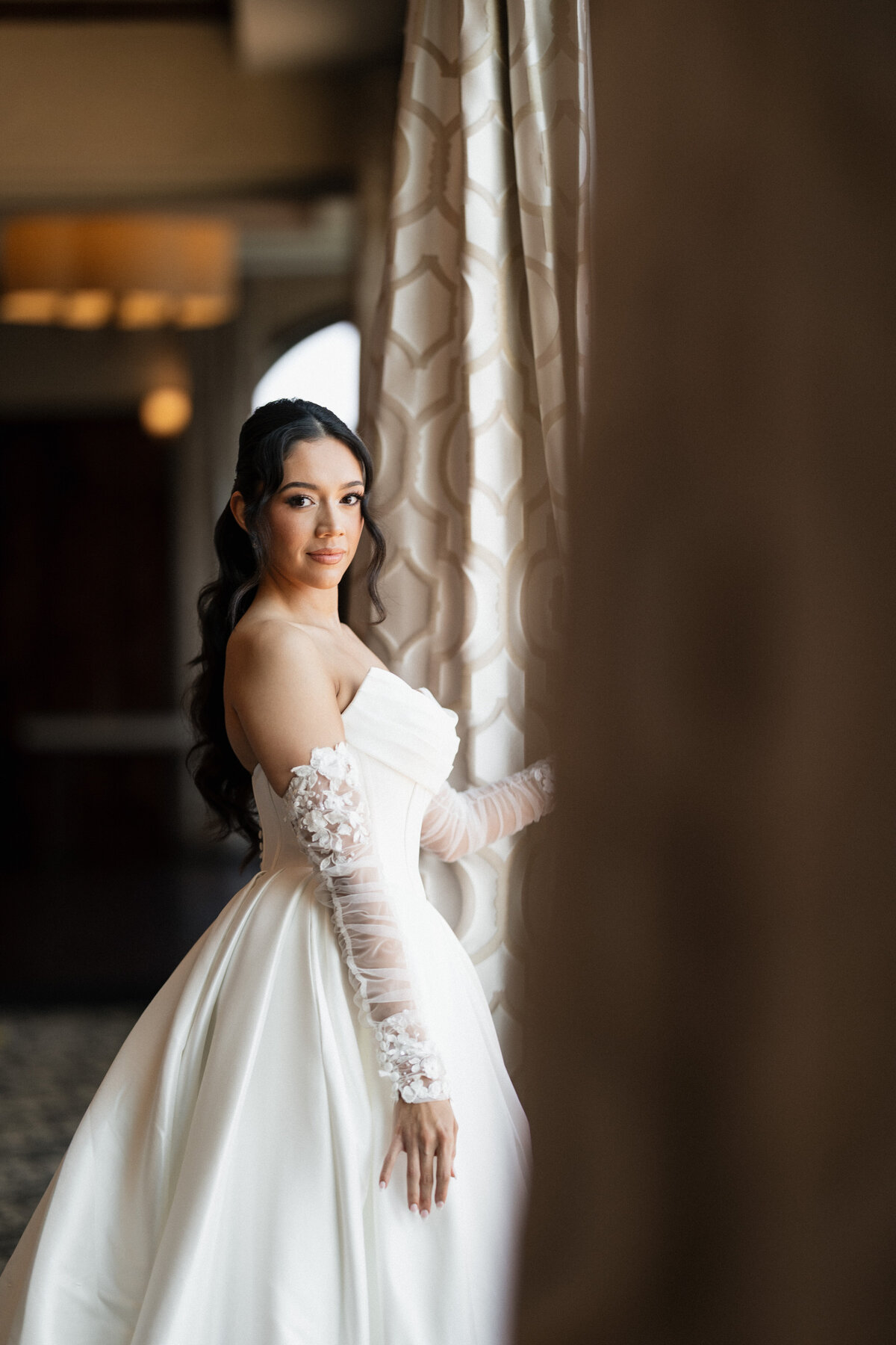 Bride standing next a window looking into the camera as she gets ready for her wedding ceremony.