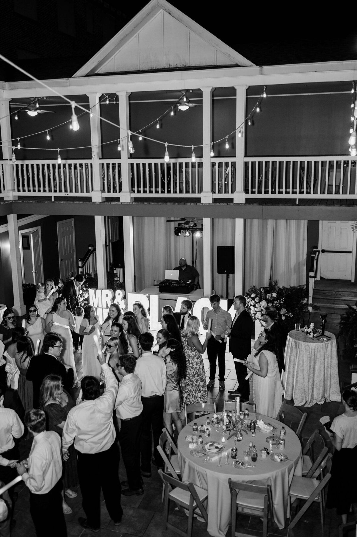 Black and white photo of a wedding reception outdoors at night; guests are dancing in front of illuminated “MR and MRS” letters, with tables, string lights, and a DJ on a balcony above the dance floor.
