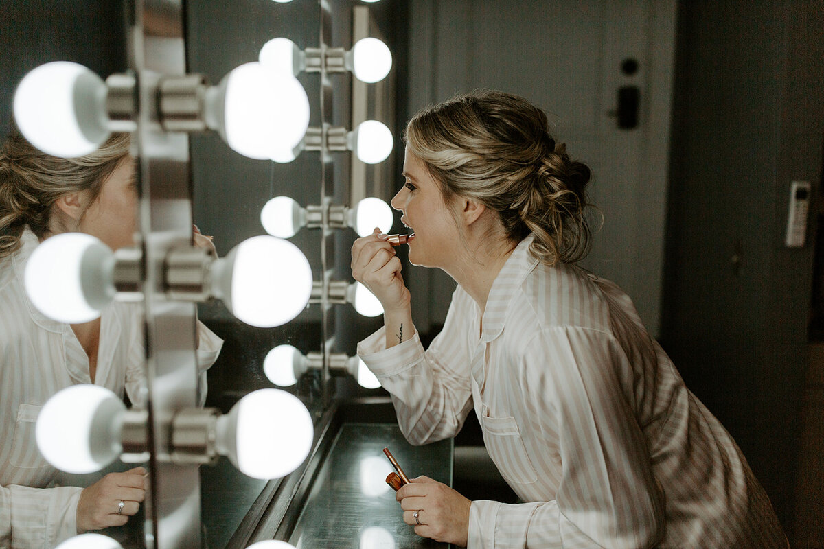 bride in stiped silk pajamas applying lipstick in a lighted make-up mirror in the preparation suite at Willowbrook Wedding venue