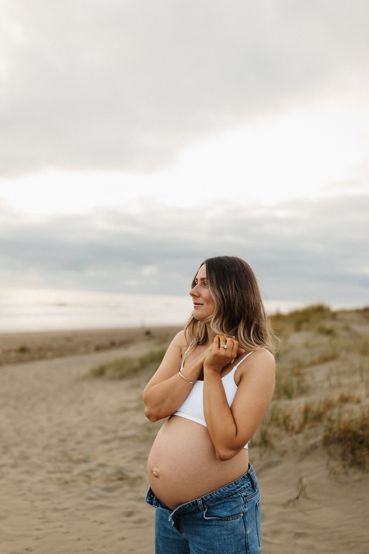 A pregnant woman holds her hands to her chest and wears a white tank top and jeans during a pregnancy photoshoot in christchurch with lucy jane photography