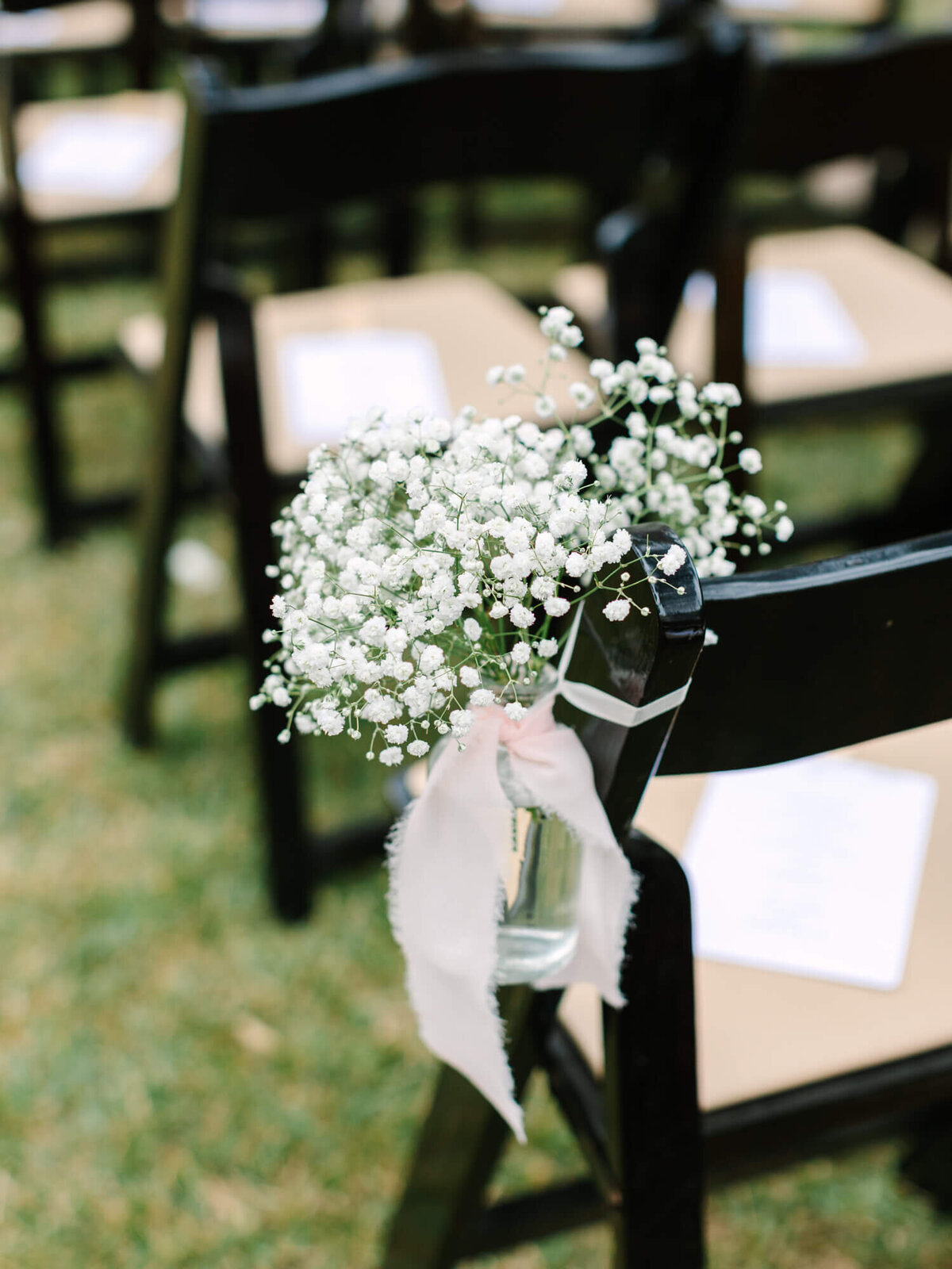 Black folding chairs are arranged outdoors on grass, with baby's breath flowers tied to one chair with a pink ribbon, creating an elegant and serene atmosphere.