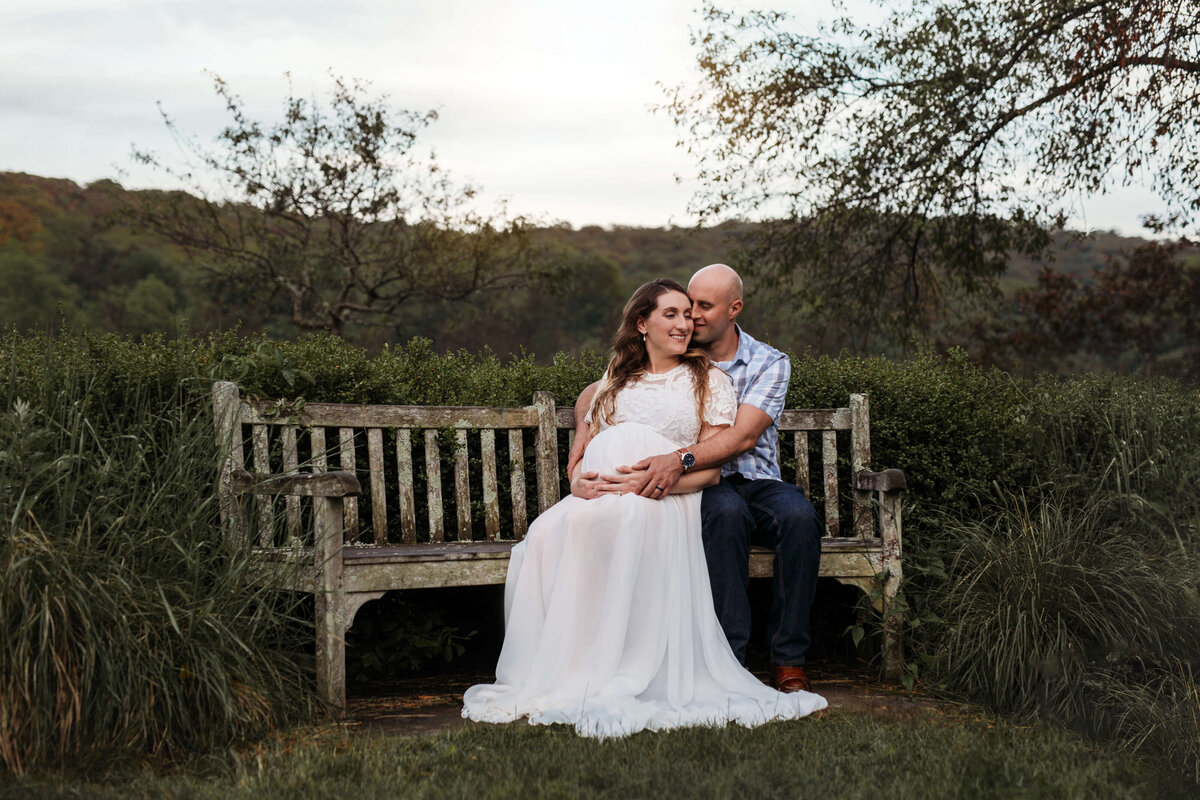 husband and his pregnant wife sitting on a bench in a garden at their outdoor maternity session
