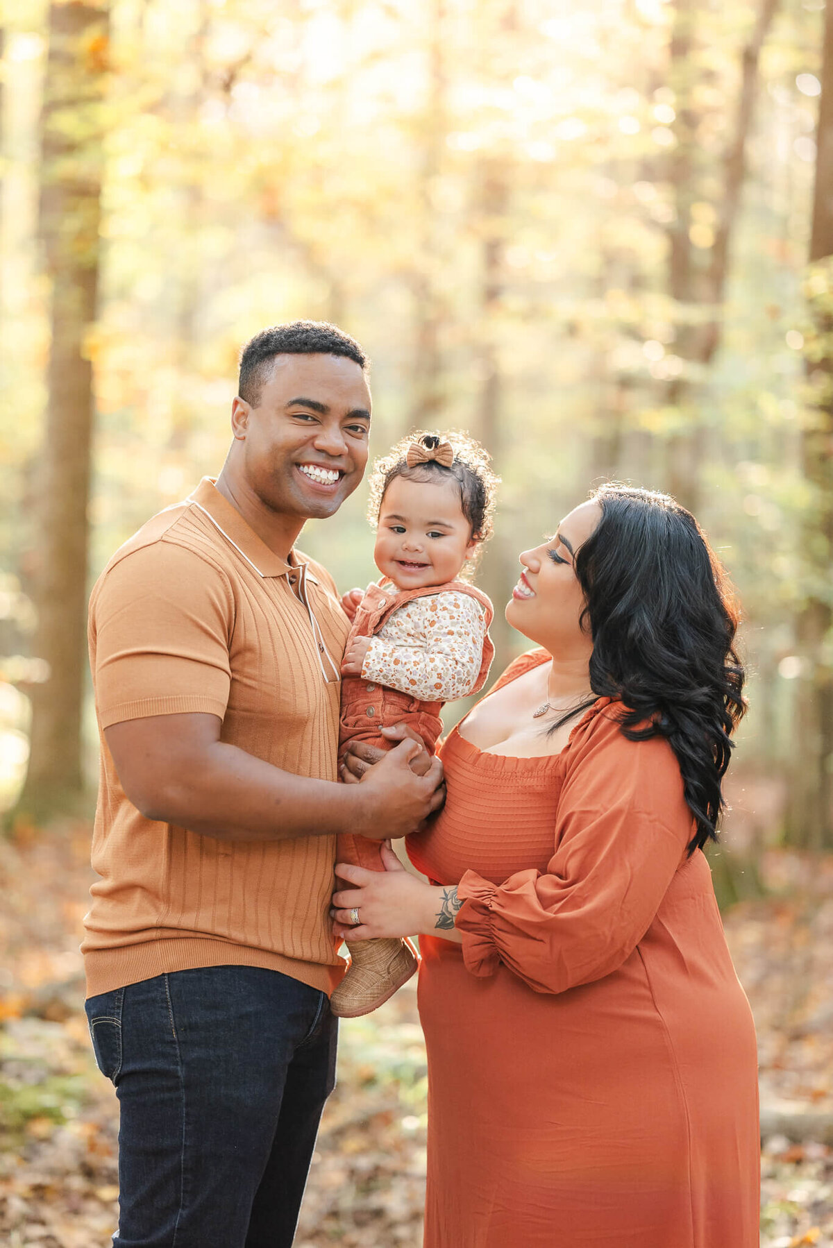A family of three poses for fall family photos. The dad is in jeans and an orange shirt. The toddler girl wears orange overalls and matching bow. The mom wears an orange dress with long sleeves.