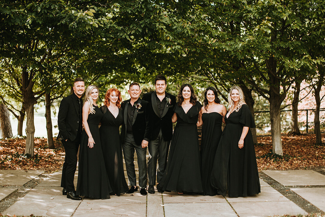 Two grooms wearing black tuxedos pose outdoors with their wedding party, who are also wearing all black.