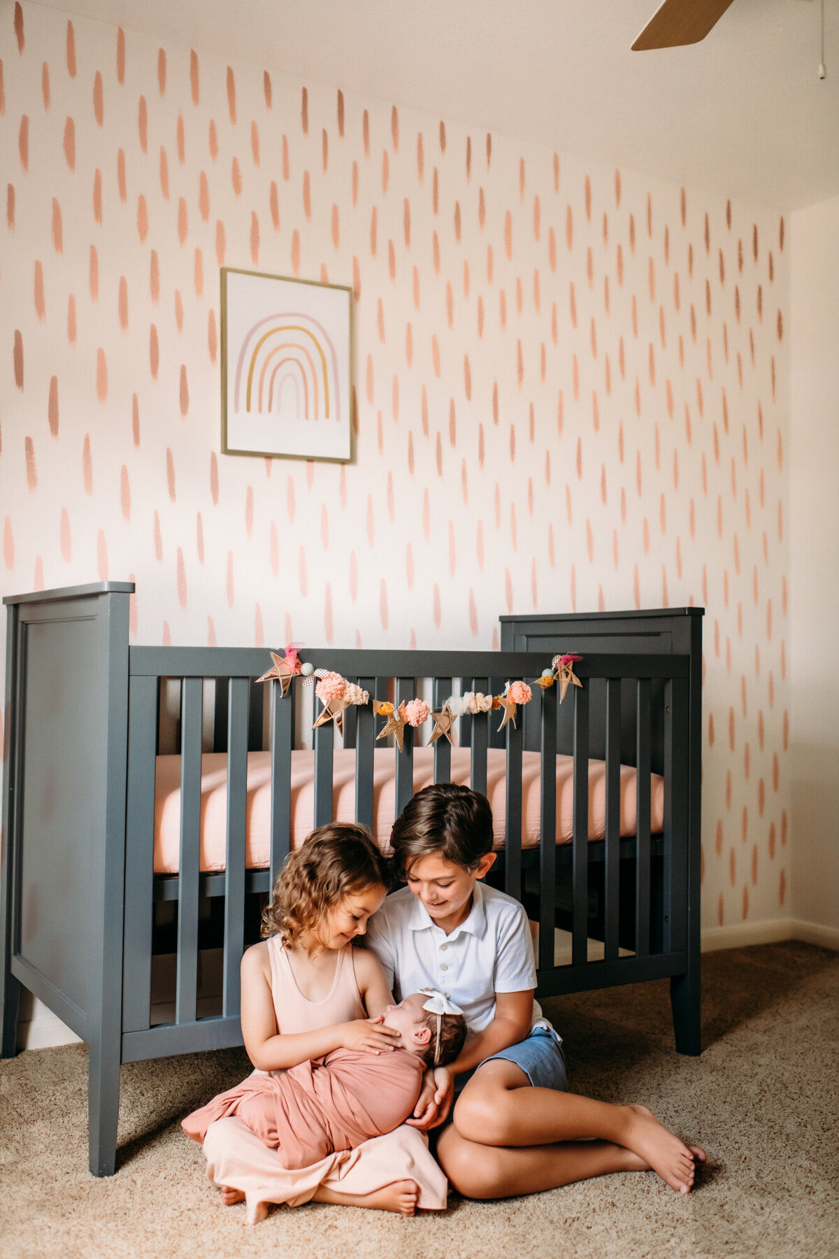 Newborn Photographer, Little boy and girl sitting in front of a crib on the floor while holding their baby sister.