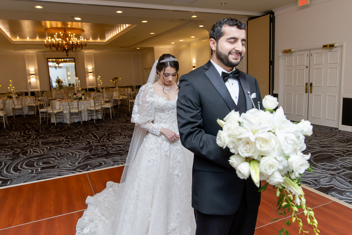 A bride stands behind her groom to do a first look. He is facing towards the camera, holding her wedding bouquet.