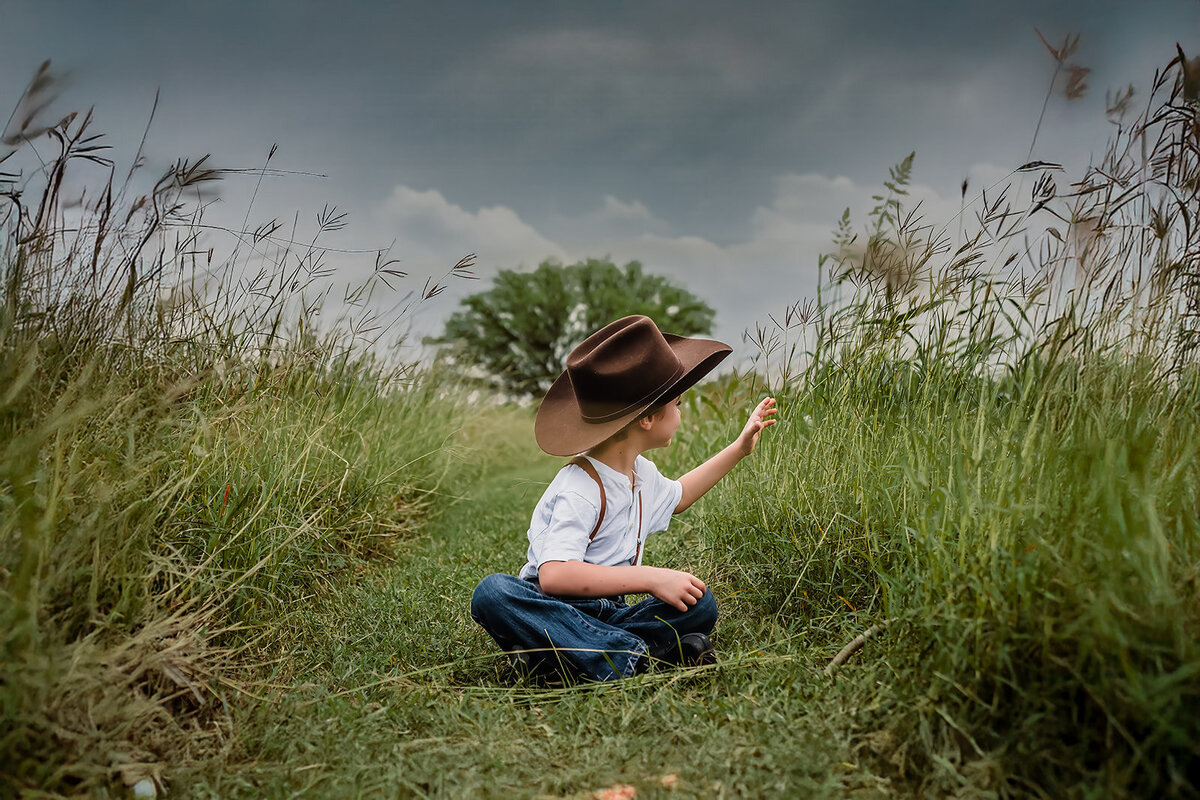 Little boy in cowboy hat sitting in field picking grass during lifestyle session in New Braunfels, Texas