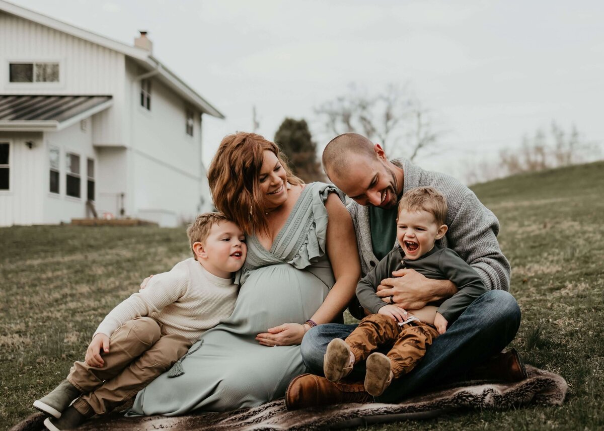 A family sits on a blanket in front of a house during a maternity session with their Pittsburgh maternity photographer.