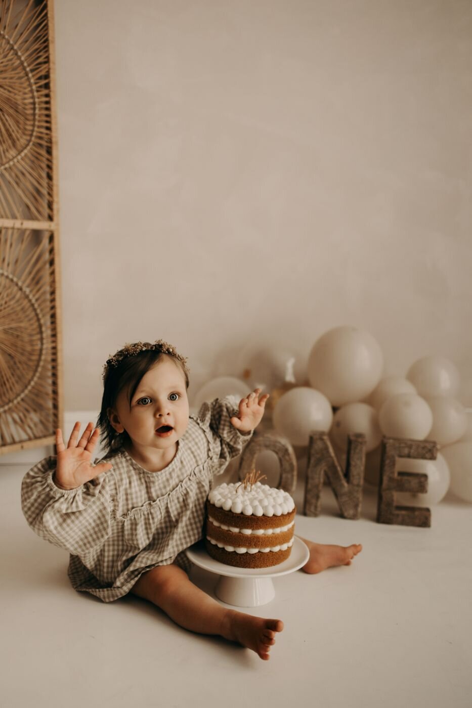 Baby girl celebrating first birthday with a cake and balloons, arms raised in excitement and surprise.
