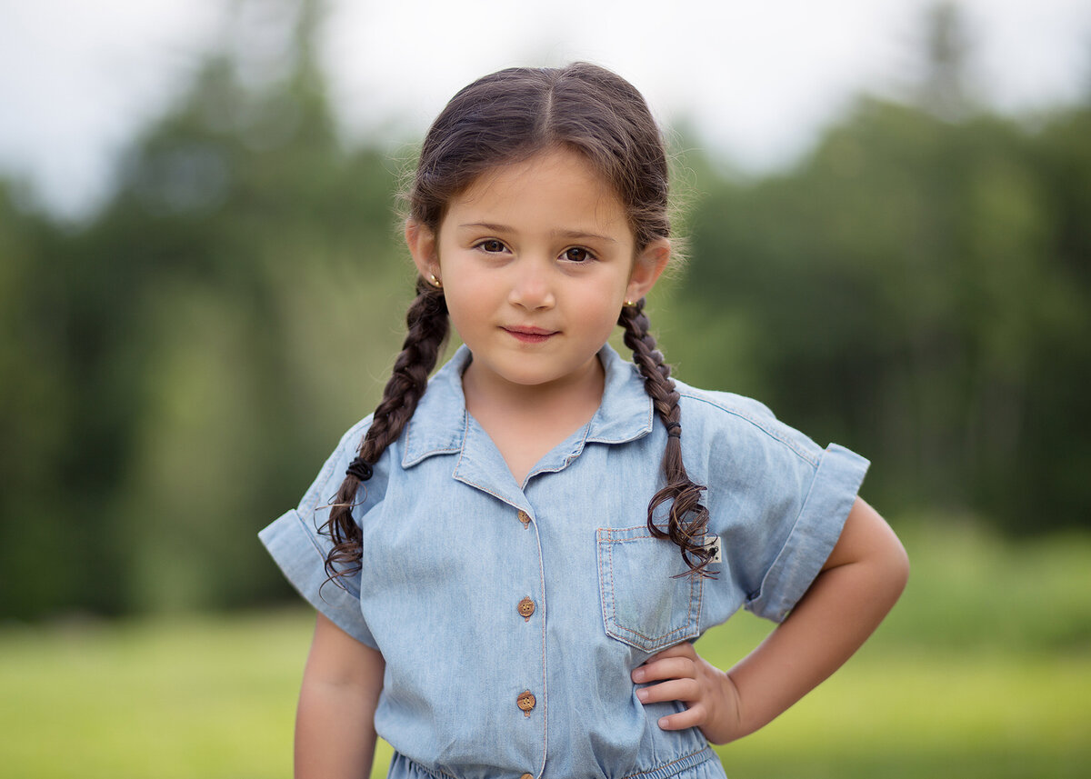 Little girls poses with her hand on her hip for a family photoshoot at a brooklyn park. Her hair is braided in french braid pigtails and she is looking at the camera with a closed mouth smile. Captured by brooklyn family photographer Chaya Bornstein.