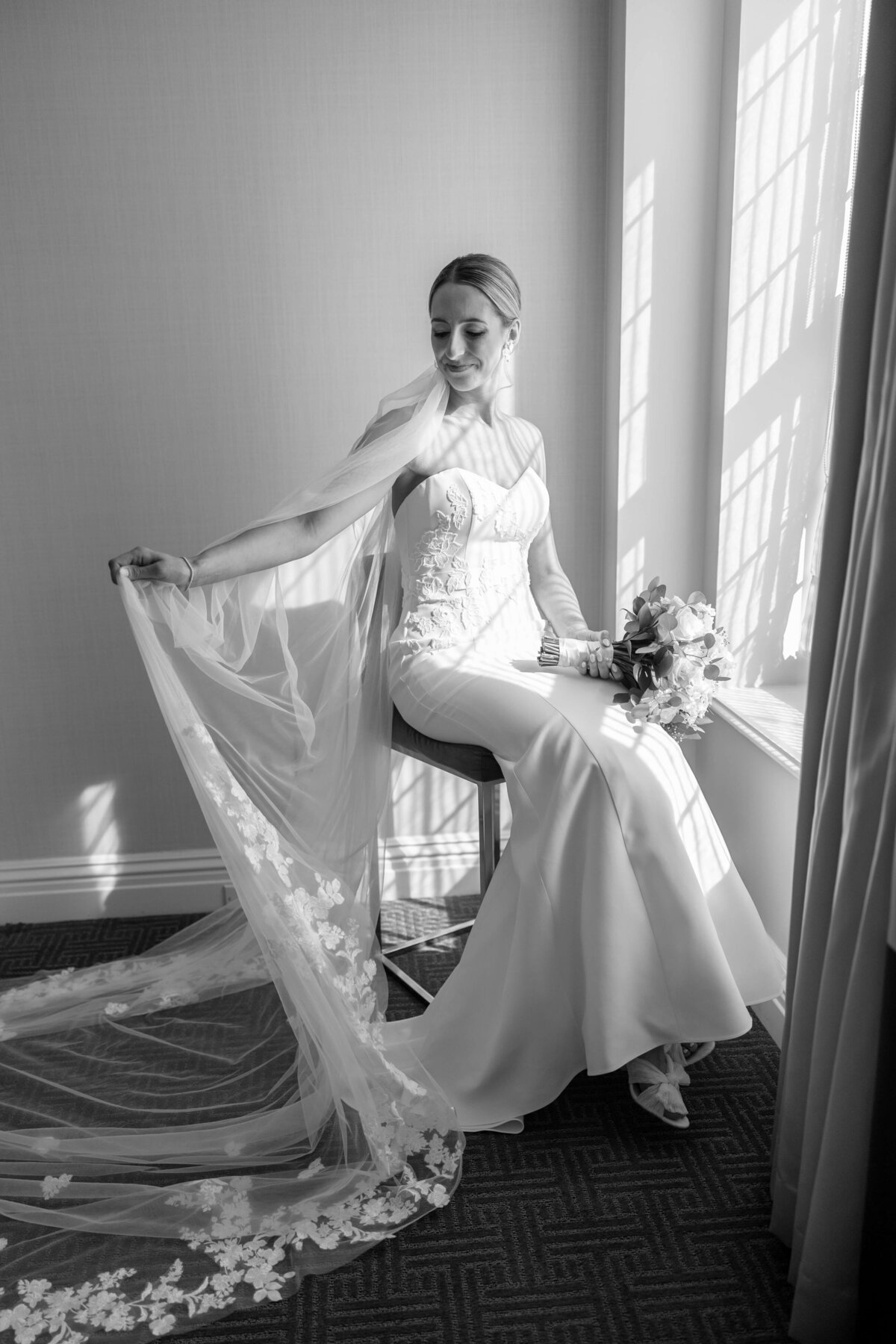 Black and white photo of a bride sitting on a windowsill. She wears a long, elegant gown with floral lace details on the train and holds a bouquet. Sunlight streams through the window, casting soft shadows on the wall.
