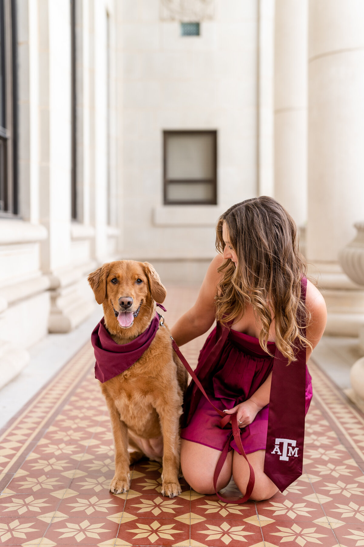 Texas A&M senior girl kneeling on ground and looking at her dog while dog smiles at camera at Administration Building columns
