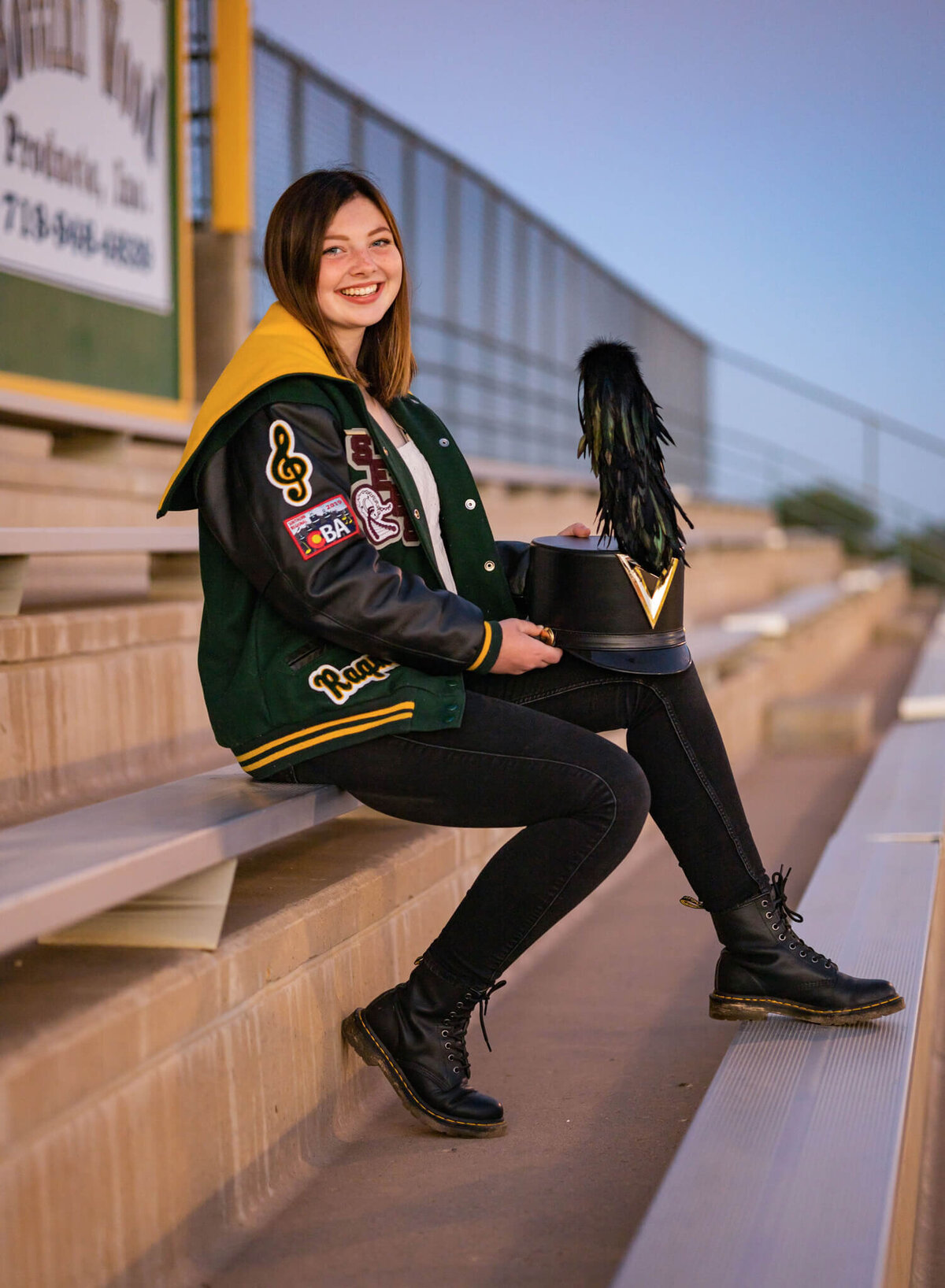 high school senior wearing her letterman jacket and holding her marching band hat sitting in the football stands