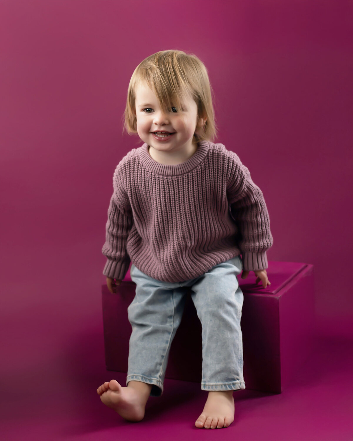 Child in a sweater and jeans sitting on a pink block with a pink background.