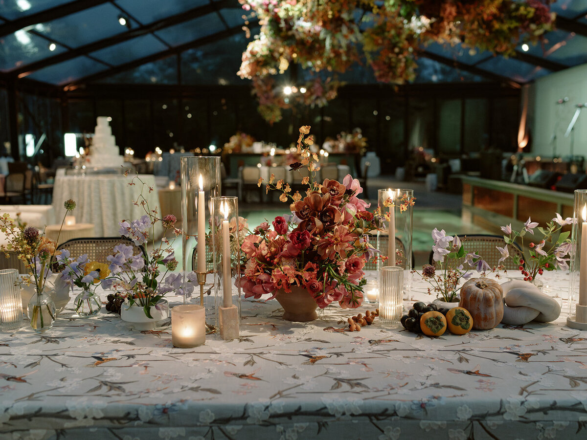 Clustered and styled floral arrangements, fruits, and art pieces for fall wedding reception. Low centerpieces mixed with ikebana arrangements and bud vases decorate the reception tables at this fall wedding. Floral centerpieces featuring orchids, ranunculus, sweet peas, and bittersweet. Styled wedding tables with fruits, curated candles, and floral arrangements. Tasteful fall color palette of copper, ruby, gold, burgundy, rust, and lavender. Design by Rosemary & Finch Floral Design in Nashville, TN.