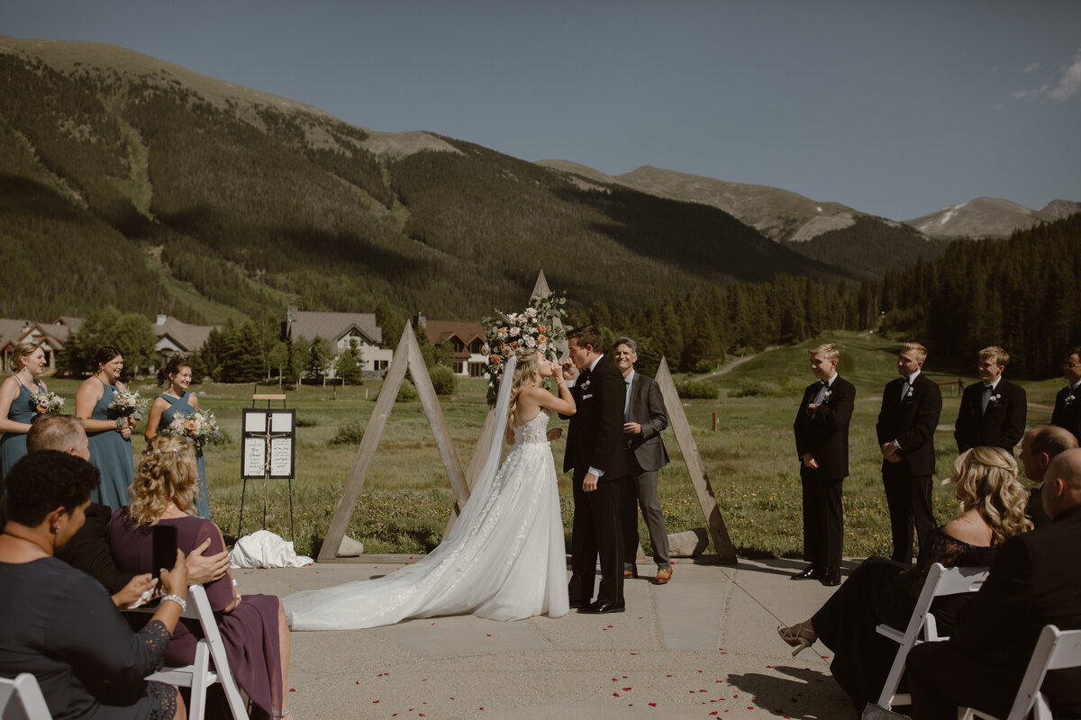 Bride and groom share a kiss during their outdoor wedding ceremony in Breckenridge, Colorado, with dramatic mountain peaks and pine forests in the background.