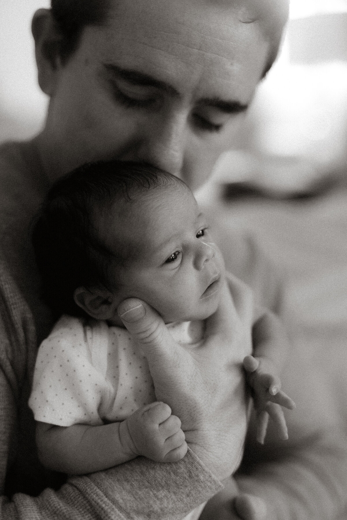 Father holding his newborn close, gently kissing the baby's head in a quiet, tender moment captured in black and white.