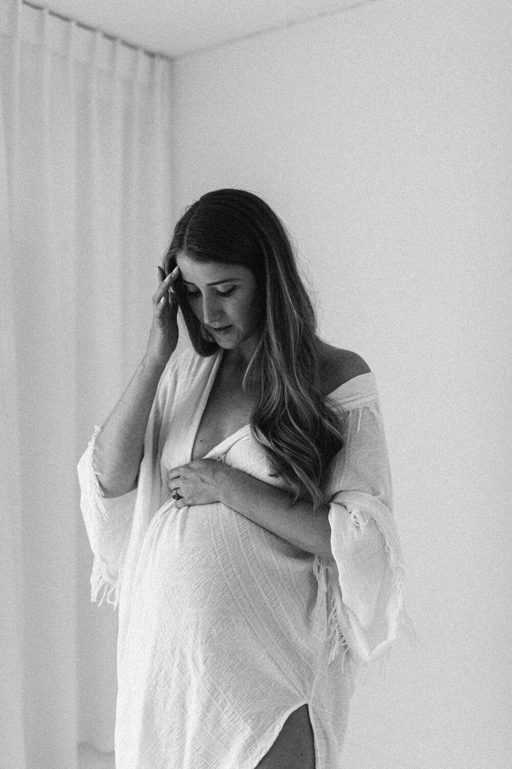 Black and white photograph of woman wearing a bedsheet who is pregnant, she is holding her belly and tucking her hair behind her ear