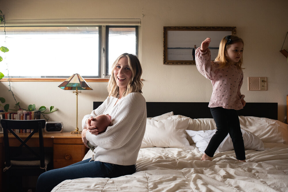 Mom holding baby on the bed while her daughter jumps on the bed behind her
