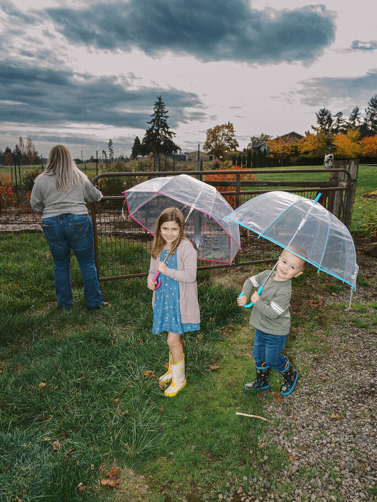 Two kids smile at the camera holding umbrellas while a woman closes a gate.