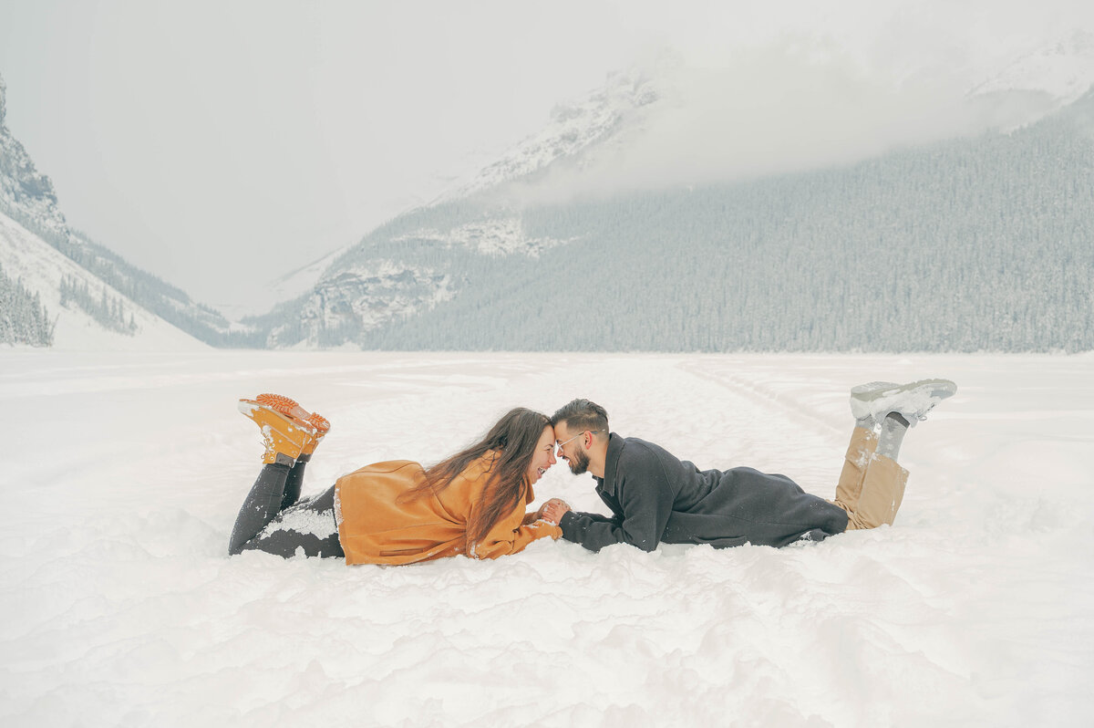 Couple lays in snow sharing a kiss during playful winter session in Banff.