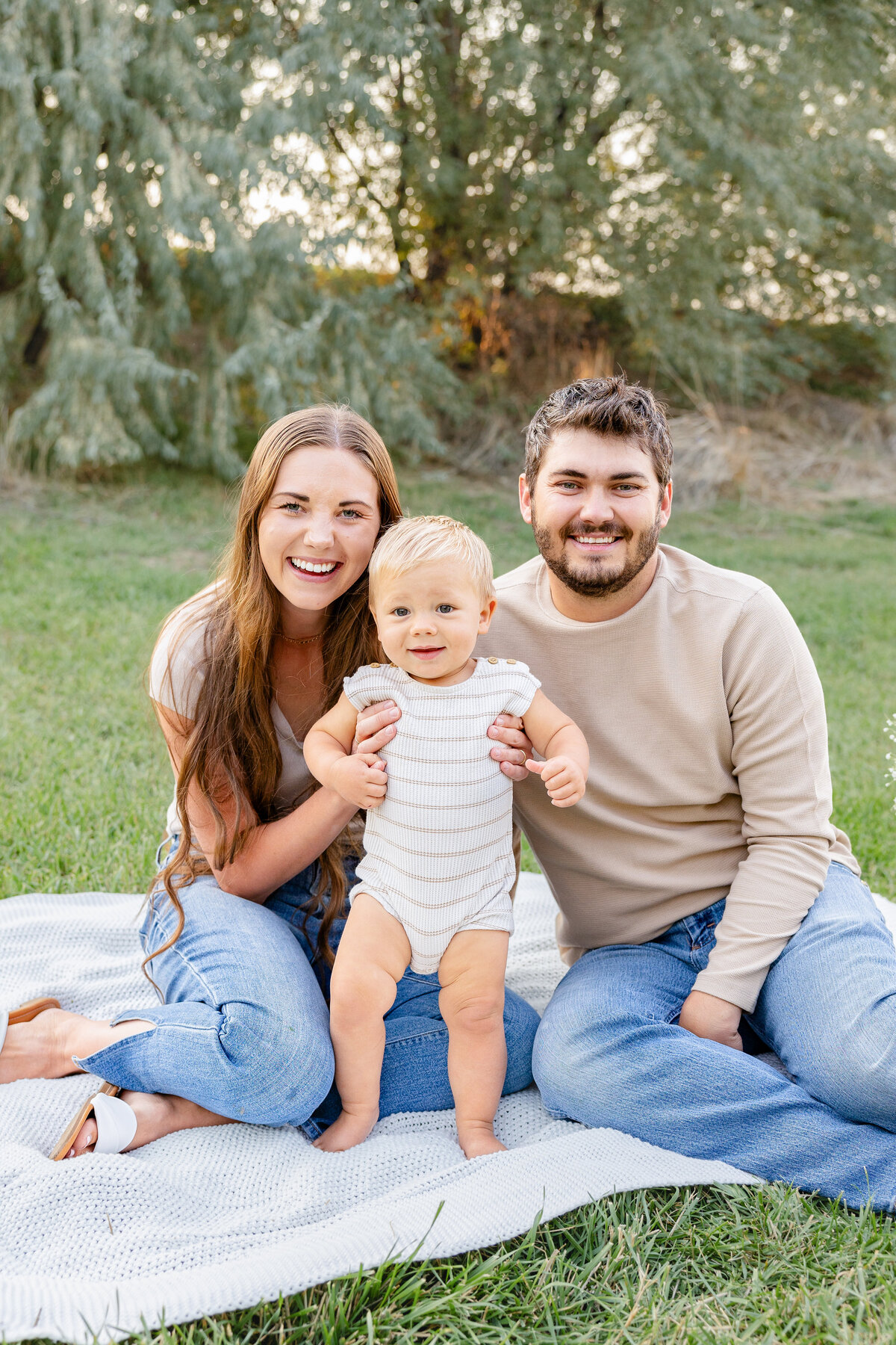 A happy young family sits on a blanket in a grassy field in Eagle Mountain, Utah. The mother and father smile as they hold their baby, capturing a moment of love and connection.