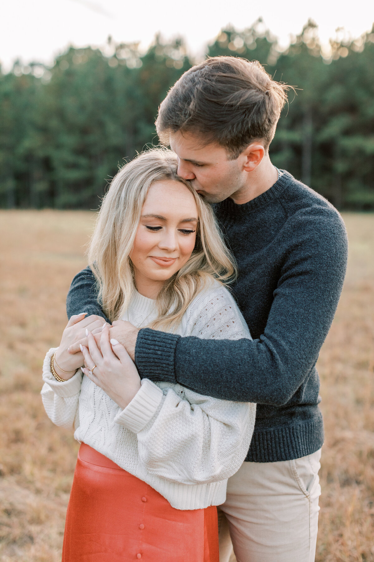 A couple embraces each other in a field surrounded by pine trees.