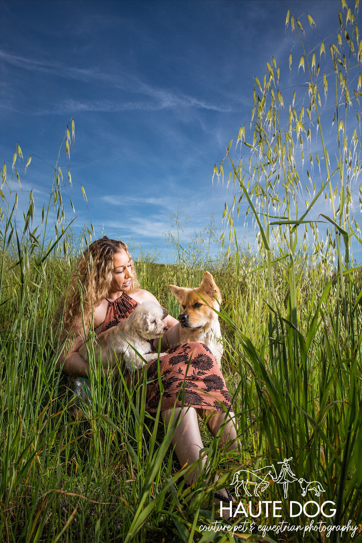 A woman sitting in a field with two dogs, surrounded by tall grass and greenery.