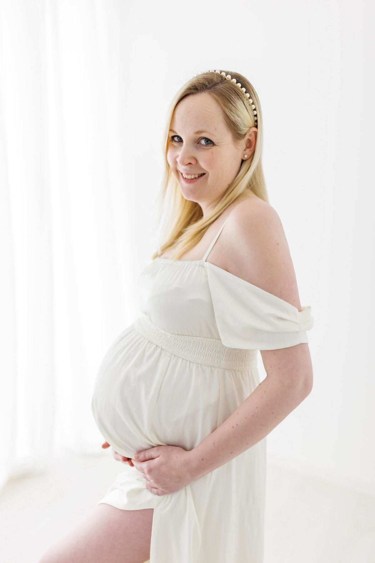 En gravid kvinne i hvit kjole holder rundt magen sin og smiler mot kamera. Hvite gardiner i bakgrunnen.