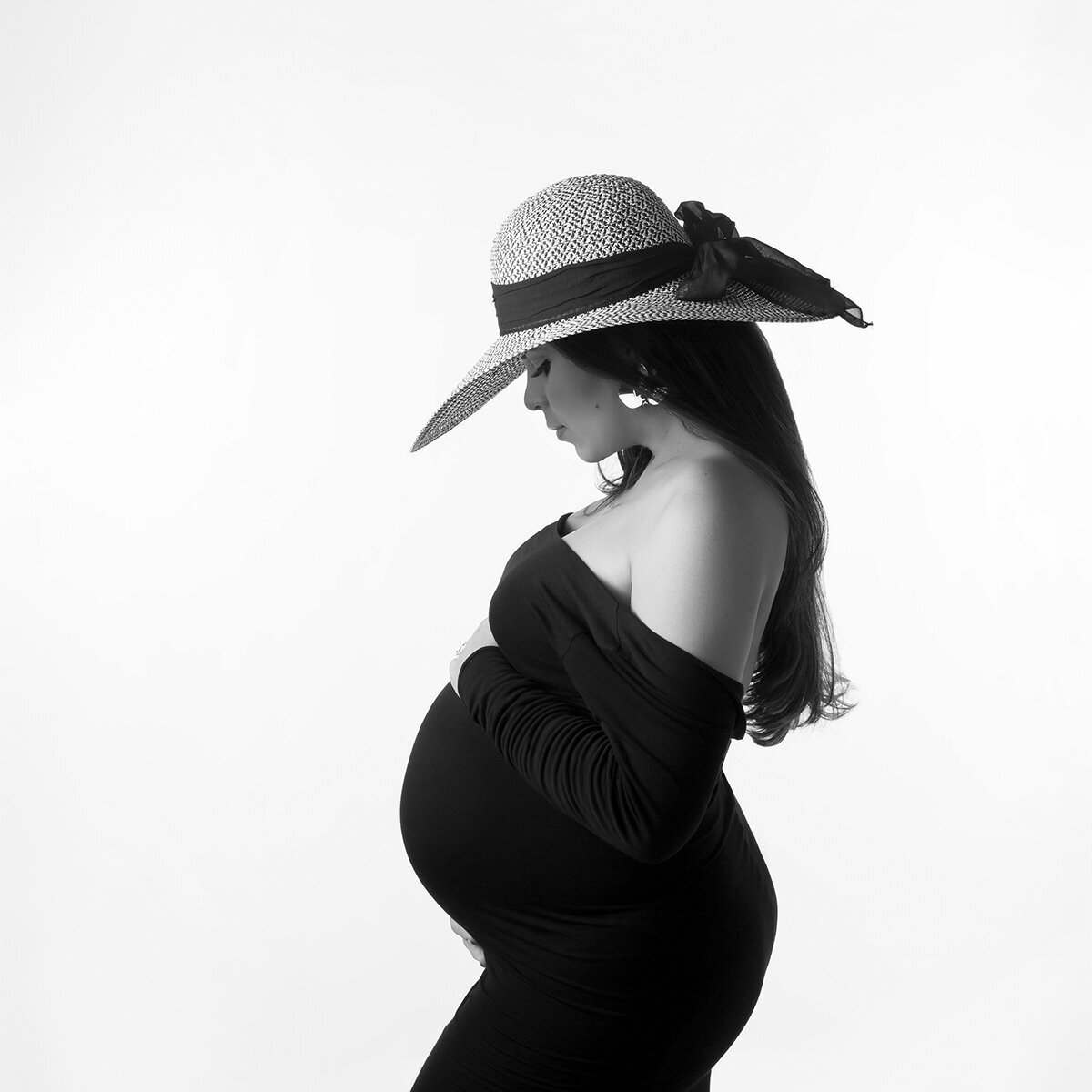 Black and white maternity photo in Charlotte, NC. A radiant Latina mom-to-be, glowing with anticipation wearing a black dress and hat, posing against a simple white background.
