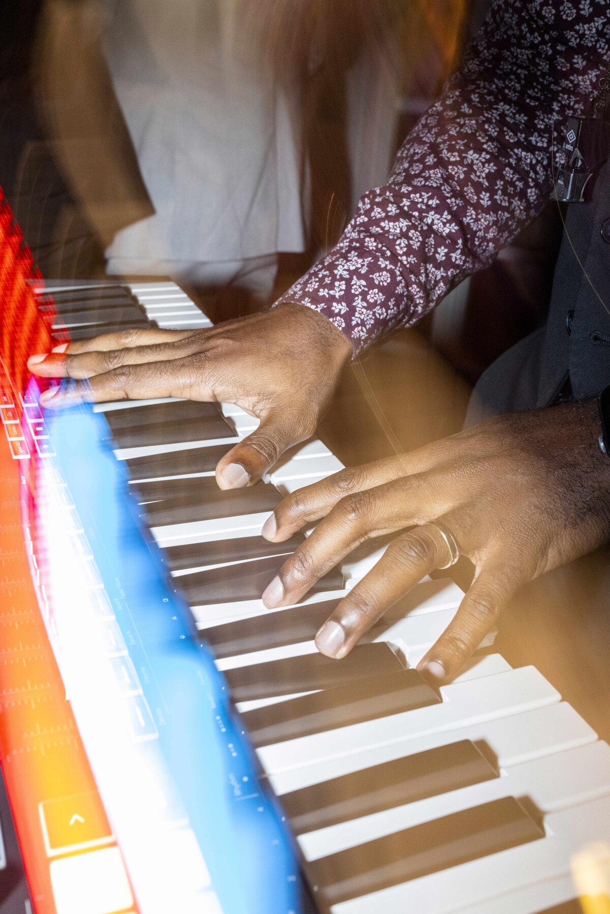 A person with a floral-patterned shirt plays a keyboard in motion, illuminated by colorful lighting. The hands are prominently featured, showing dynamic movement and energy.
