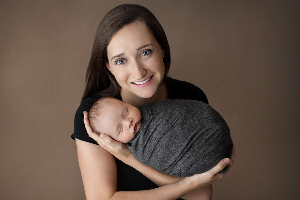 new mother smiling holding her newborn baby boy on a brown backdrop in a newborn session