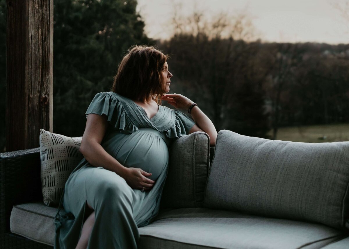 A pregnant woman sitting on a couch in Pittsburgh in a green dress is captured by the maternity photographer.