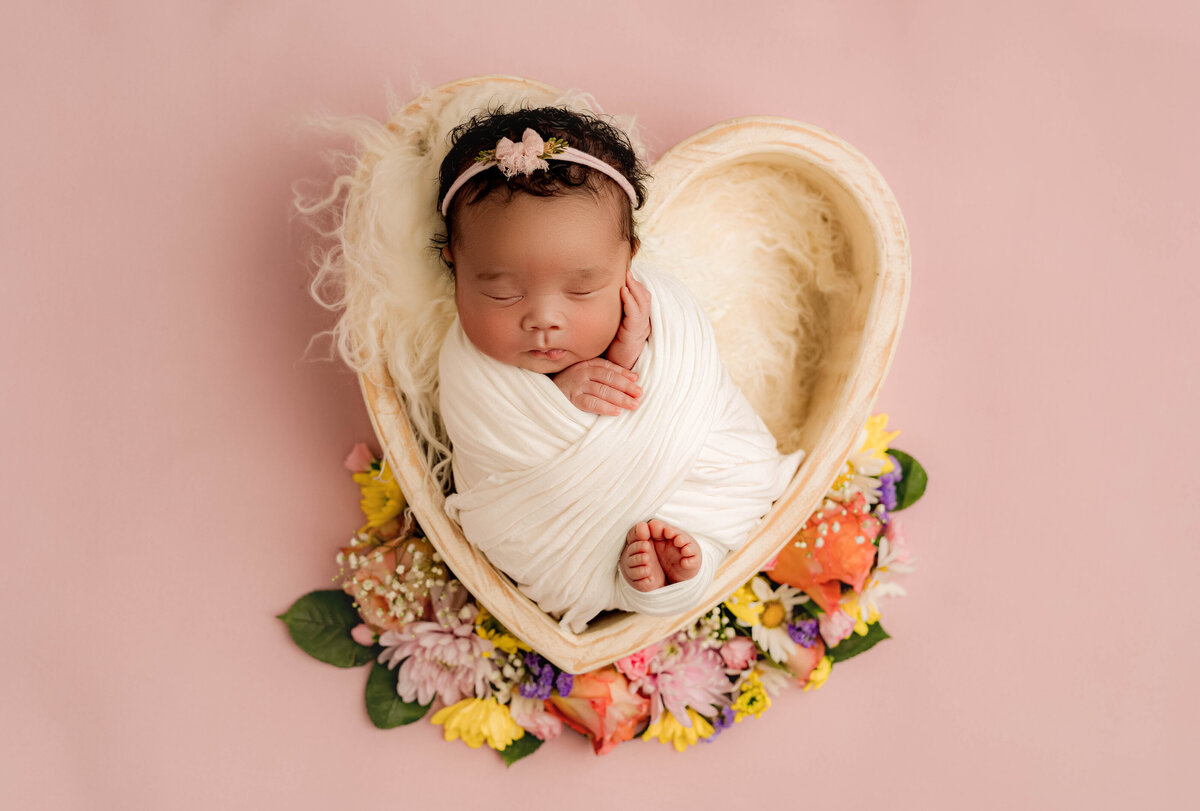 In a serene Charlotte newborn photography studio, a baby sleeps soundly wrapped in a white blanket on a heart-shaped bed lined with soft fur. The pink backdrop and colorful flowers add charm, while the floral headband completes the peaceful scene.