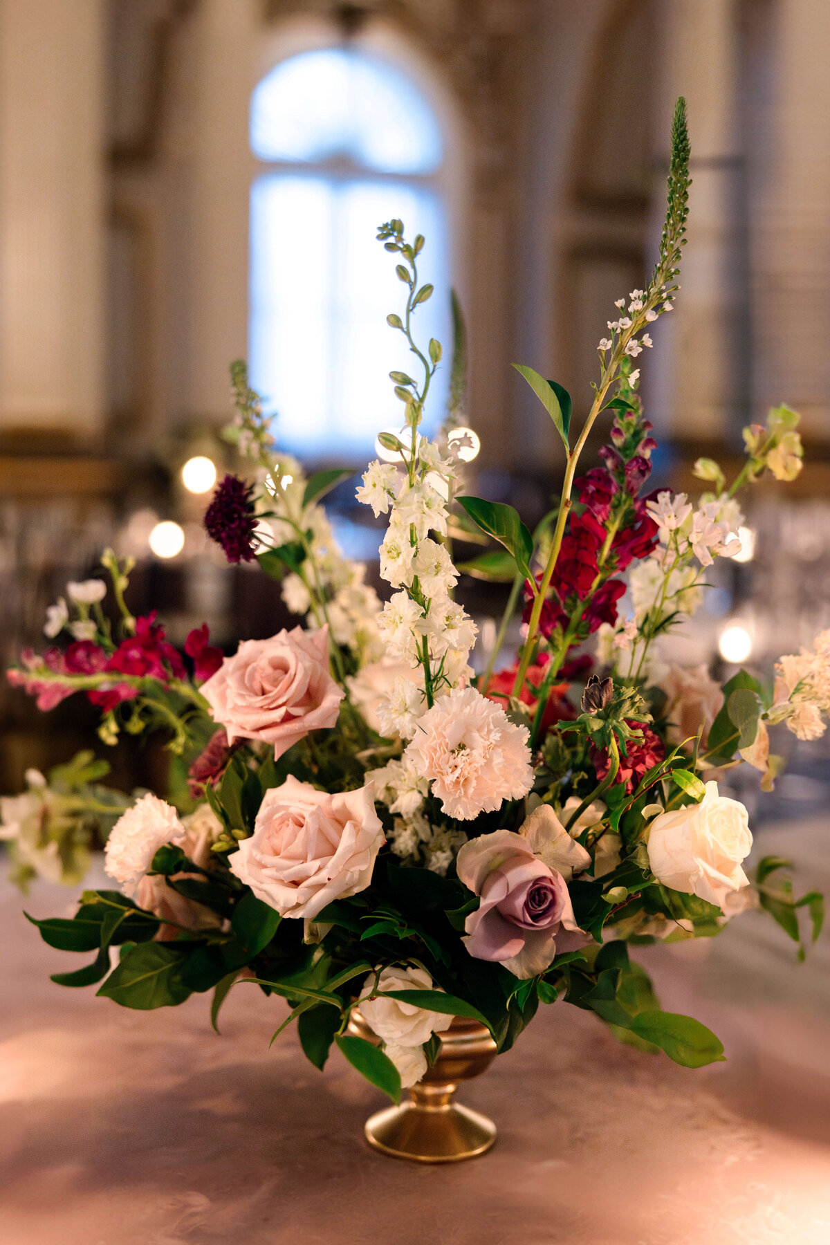 A floral arrangement in a gold vase featuring pink roses, white and purple flowers, and green foliage. The background is a softly lit, elegant room with arched windows.