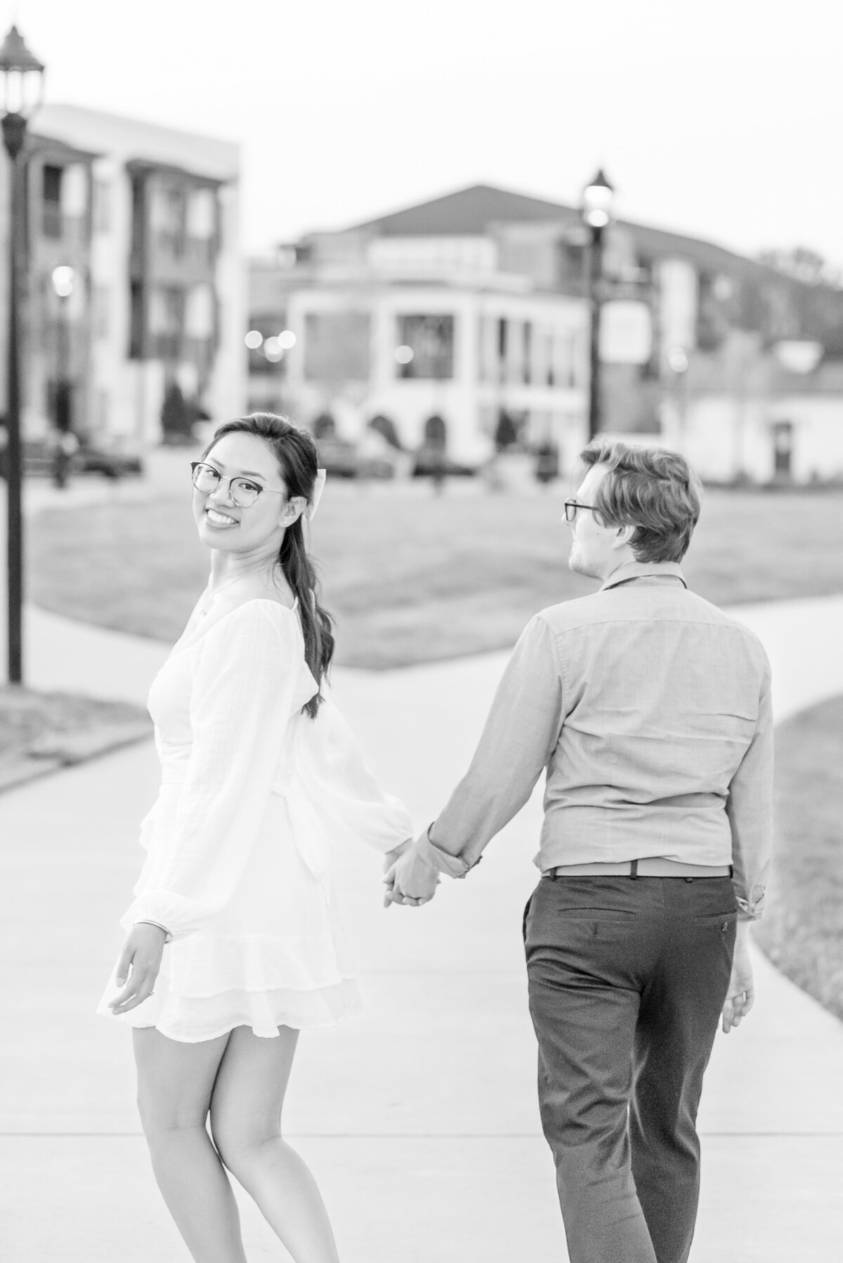 Black-and-white-image-of-an-engaged-couple-walking-away-while-the-Asian-female-looks-back-over-her-shoulder-at-the-camera-smiling-at-the-University-City-Boardwalk-by-Charlotte-wedding-photographers-DeLong-Photography