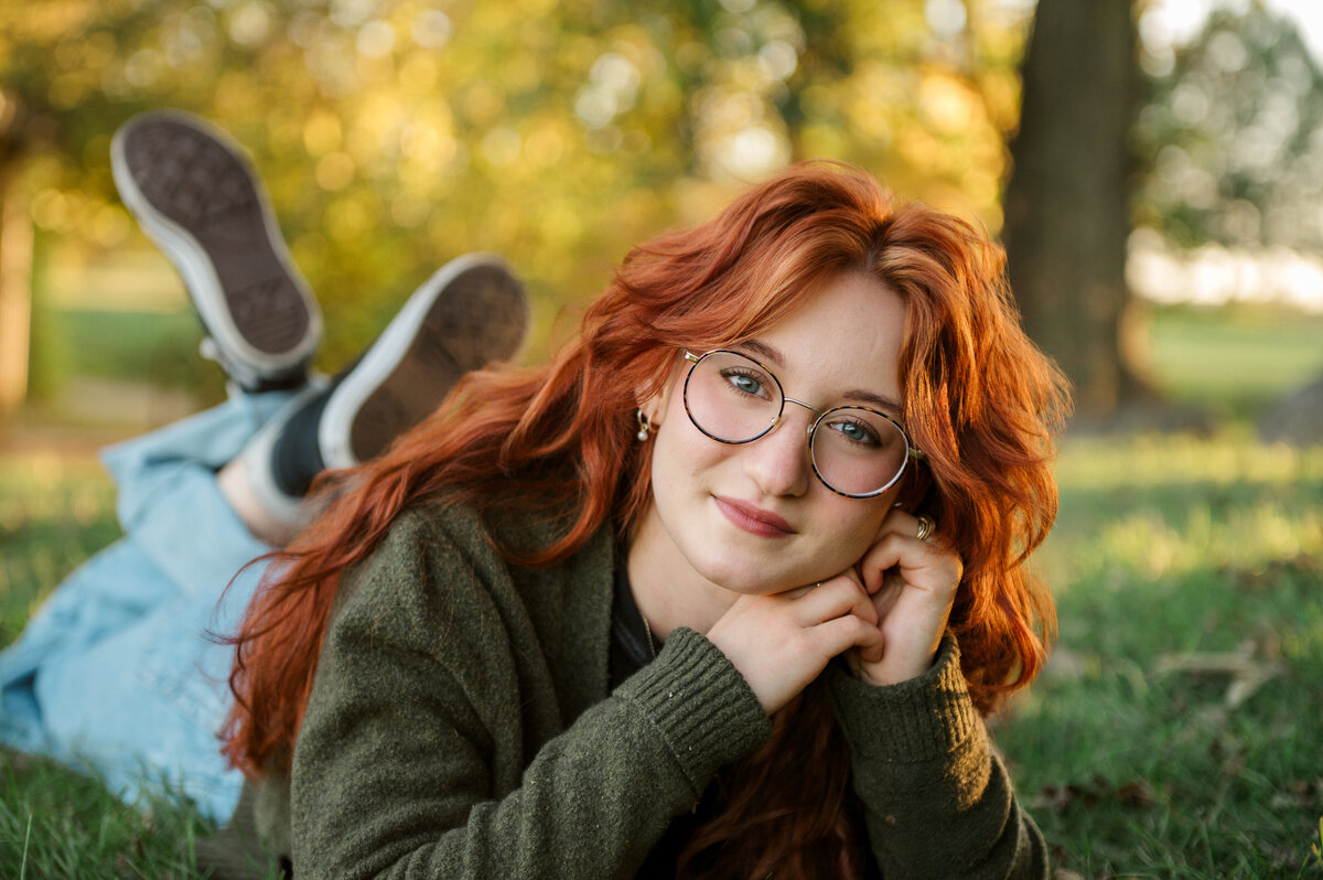 Senior girl with red hair and glasses lying in the grass during golden hour – captured by York PA senior photographer Kara Michelle Photography.