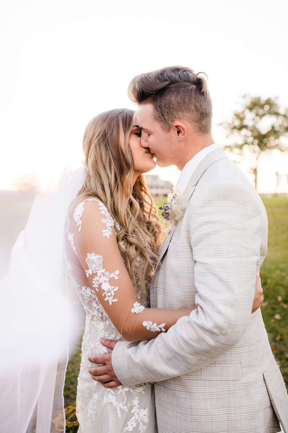 Bride in lace dress with veil flowing behind kissing groom with hands wrapped around each other